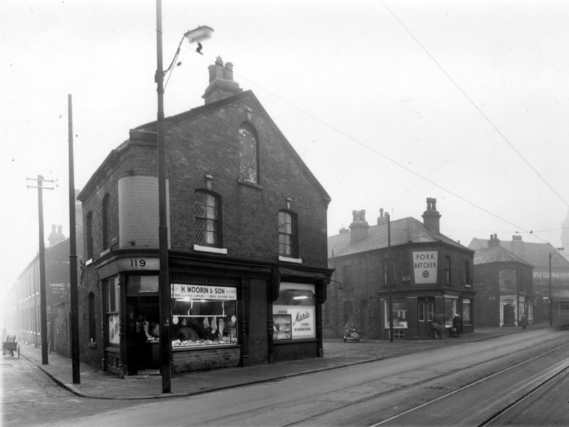 On the left a wooden cart loaded with ladders stands in Addington Street. On the corner is number 119 Dewsbury Road, the H. Moorin and Son butcher using a large set of scales. Cuts of meat are visible in the window. On the right at number 121 is the M. Kirby run Marie ladies hairdresser. Here is Addington Place with the T. Ward pork butchers at number 123 Dewsbury Road. Pictured in November 1958.