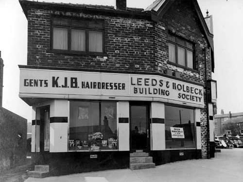 K.J.B. gents hairdressers and the offices of the Leeds and Holbeck building society on Lower Wortley Road. Both have accommodation above and an alley runs down the left hand side. Pictured in April 1936.