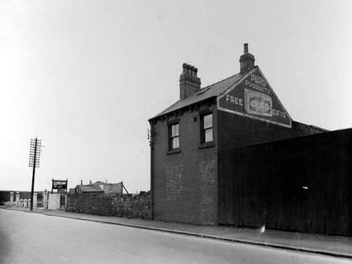 Part of the Puro Soap Co's Prospect works with a soap advertisment on the gable-end of the building. A wooden fence obscures the rest of the building and factory. There is a petrol station at the left of the soapworks that has a Cleveland and a Pirelli tyre advertising board. Pictured in April 1936.