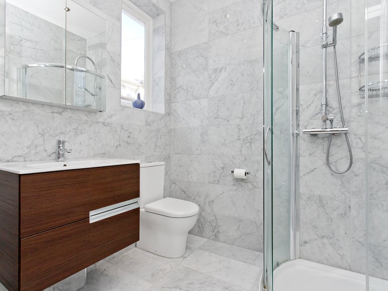 The principal bedroom’s en-suite shower room comprises a corner enclosure with a rainfall showerhead and handset, a vanity unit with a basin inset, a WC, and a mirrored, wall-mounted vanity cabinet, all enveloped by stylish marble wall and floor tiles.