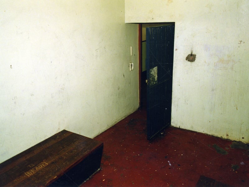 Inside a disused cell. A wooden bench is fitted against the wall with graffiti gauged into it. The name carved reads 'Brady'.