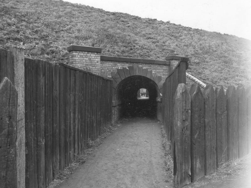 Wooden fences made from two rows of old railway sleepers are seen to the left and right of a passageway leading to a tunnel through the railway embankment. The tunnel formed part of a footpath leading from Brown Lane to Oswald Grove. Construction work looks to be going on in the yard at the other end of the tunnel. Pictured in October 1940.