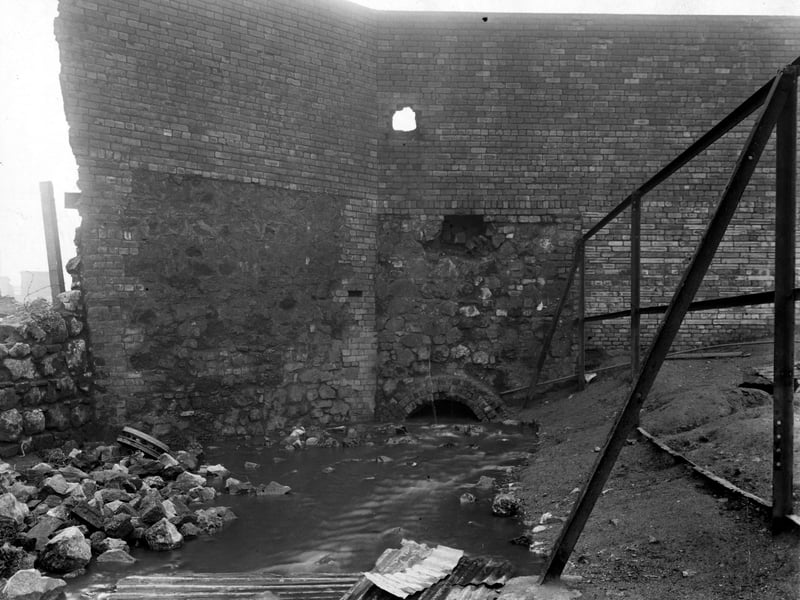 A view of Balm Beck flowing towards an opening beneath a partially demolished brick and stone wall. Sheets of corrugated iron can be seen in the foreground next to a pile of rubble in the Bower Road area. Pictured in November 1949.