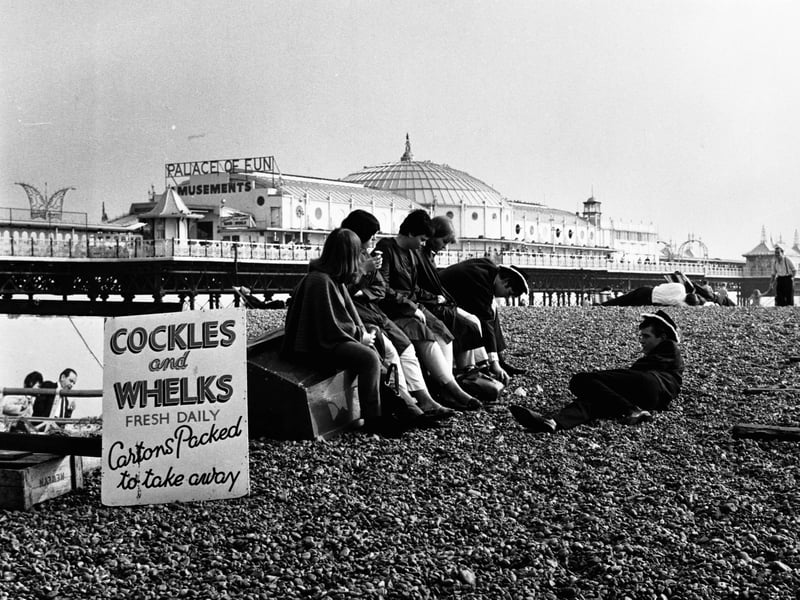 A group of people relaxing on the beach, next to a sign selling cockles and whelks, with the pier and amusement arcade behind them, at Brighton in the mid-1960s
