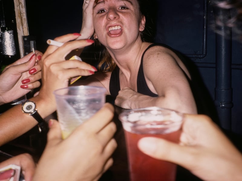 Hands holding plastic cups and a cigarette next to a woman sitting at a table in a nightclub in 1991