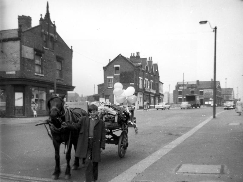 A boy holds onto the reins of a horse which is pulling a rag and bone cart. The cart is piled with old clothes and has several balloons tied to the back. View looks along Burmantofts Street. The entrance to Moody's Yard is just visible on the left edge. Numbers on Burmantofts Street run to the right in ascending order from number 15 on the left. Pictured in 1964.