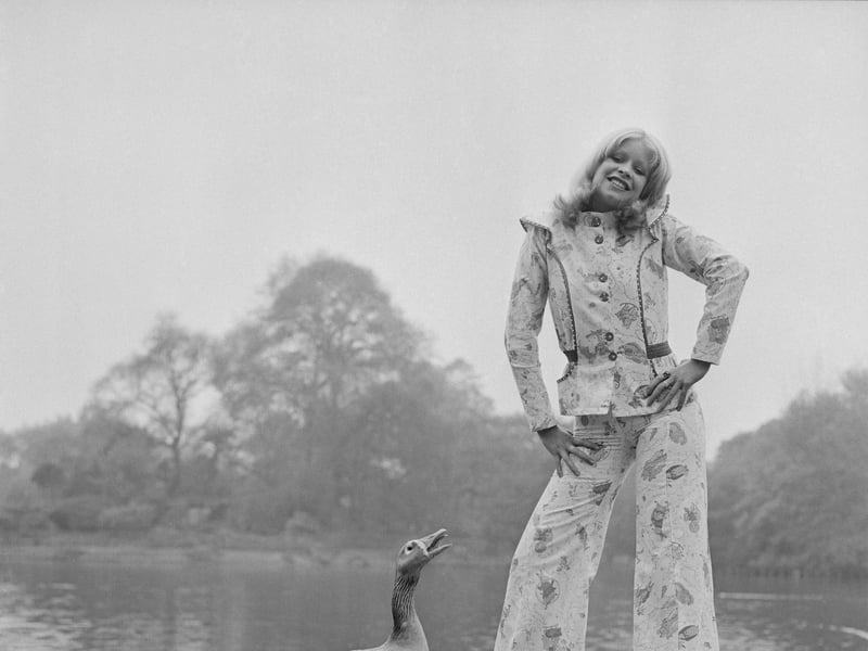 A goose looks on as a model sports a patterned pant suit, in May 1972
