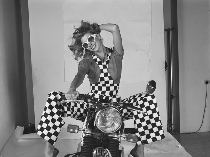 A young model wearing checkered overalls and sitting astride a motorcycle during a fashion shoot in August 1973