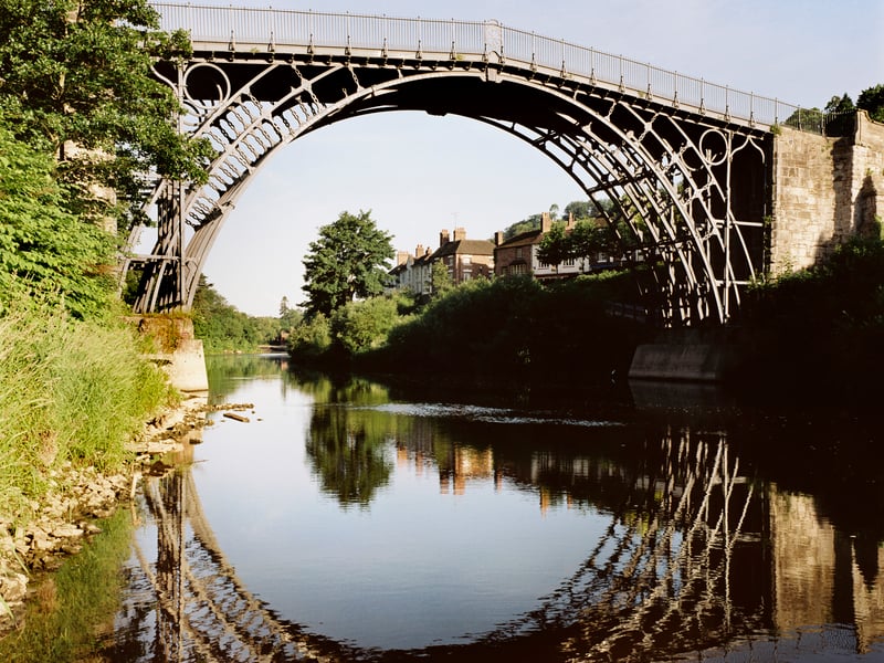 View of the bridge looking down the River Severn with reflections shown in the water. Abraham Darby established an iron works at nearby Coalbrookdale in 1708. His grandson, Abraham Darby III built this bridge, the first cast iron bridge in the world, between 1777 and 1779. It was designed by Thomas Farnolls Pritchard and opened in 1781.