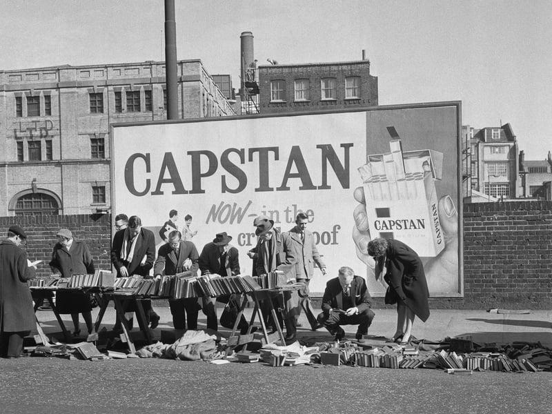 A billboard advertising Capstan cigarettes is the backdrop to builders browsing the books on sale at a bookseller's stall in March 1961