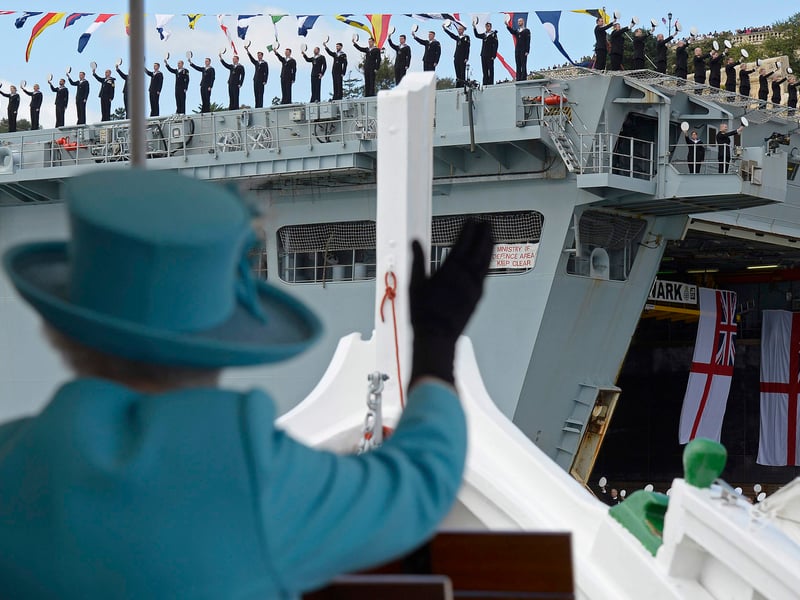 Queen Elizabeth II waves to British Royal Navy crew members performing a salute on the HMS Bulwark amphibious assault ship during a tour of the Grand Harbour in a traditional Maltese fishing boat, with the HMS Bulwark amphibious assault ship seen in the background on November 28, 2015 in Valletta, Malta.