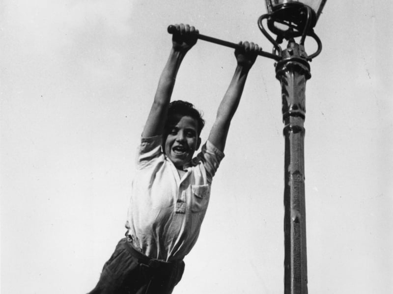 A boy swings from a lamppost in a 'play street', barred to through traffic to make it safe for children to play, in 1955