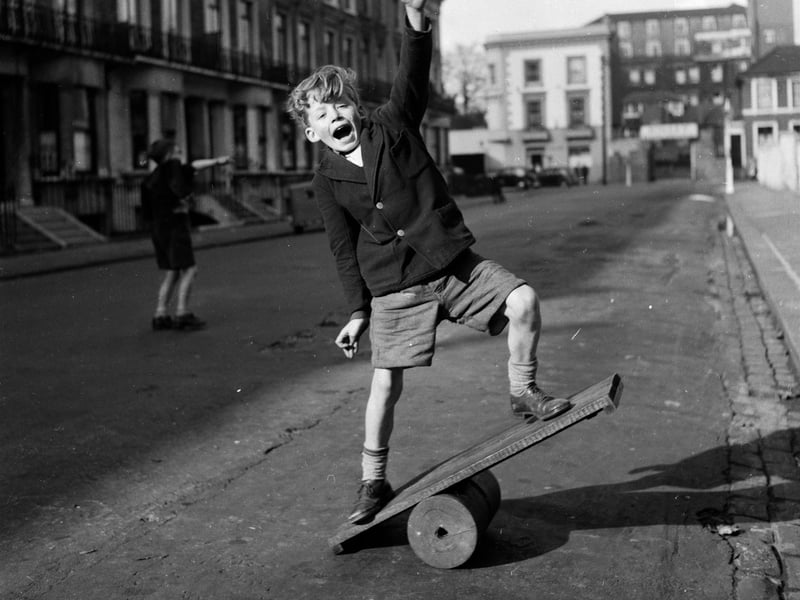 A boy tests his balance on a wiggle board, constructed from a wooden board positioned on a cylindrical roller, in June 1956