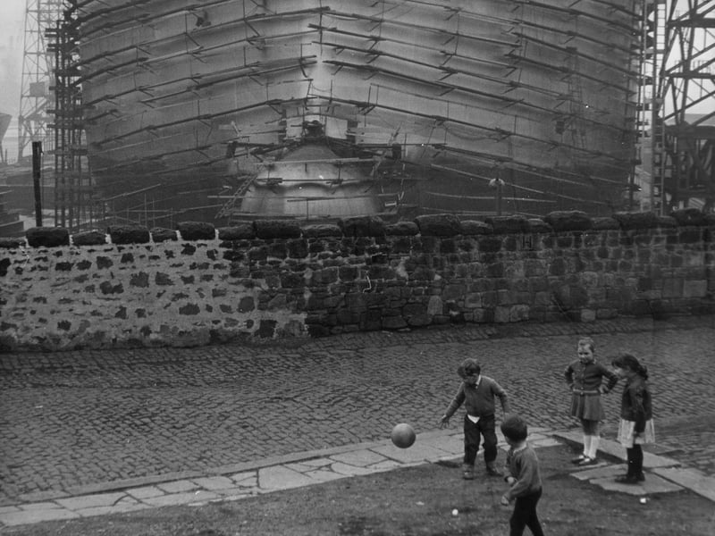 Children play a the bottom of Gerald Street, Wallsend-on-Tyne, in the shadow of the hulking tanker, 'Narica', being built in the dockyard beyond the street wall, in 1967