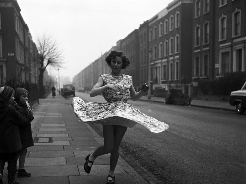 A girl twirling her dress in the street as two younger children look on, in February 1958