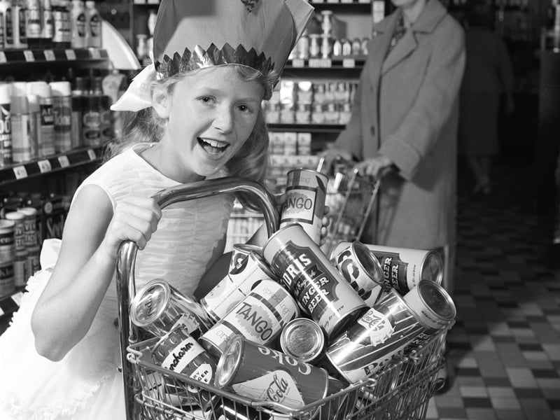 A little girl in a party outfit wheels a trolley full of canned soft drinks at a supermarket in July 1963