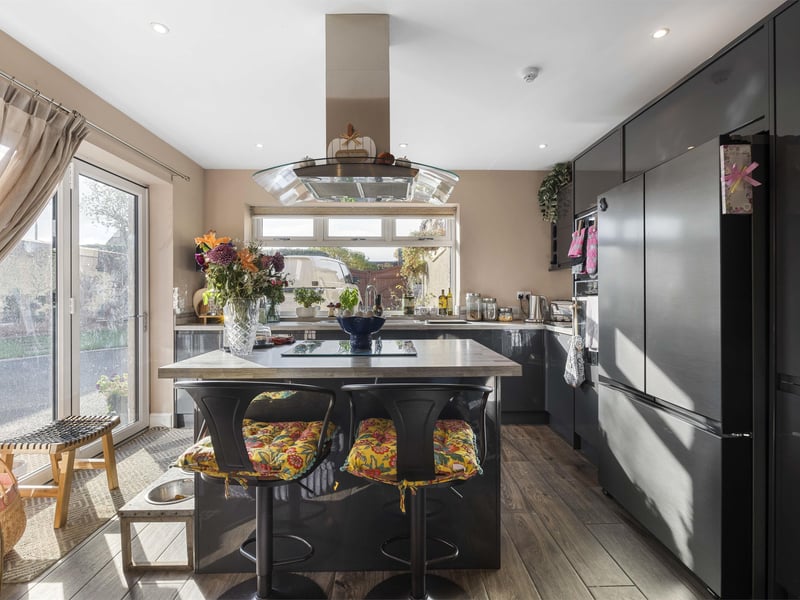 The open-plan kitchen and dining area create a stylish modern living space, featuring a sleek glossy grey fitted kitchenette with an island breakfast bar, electric hob, and integrated overhead extractor. 