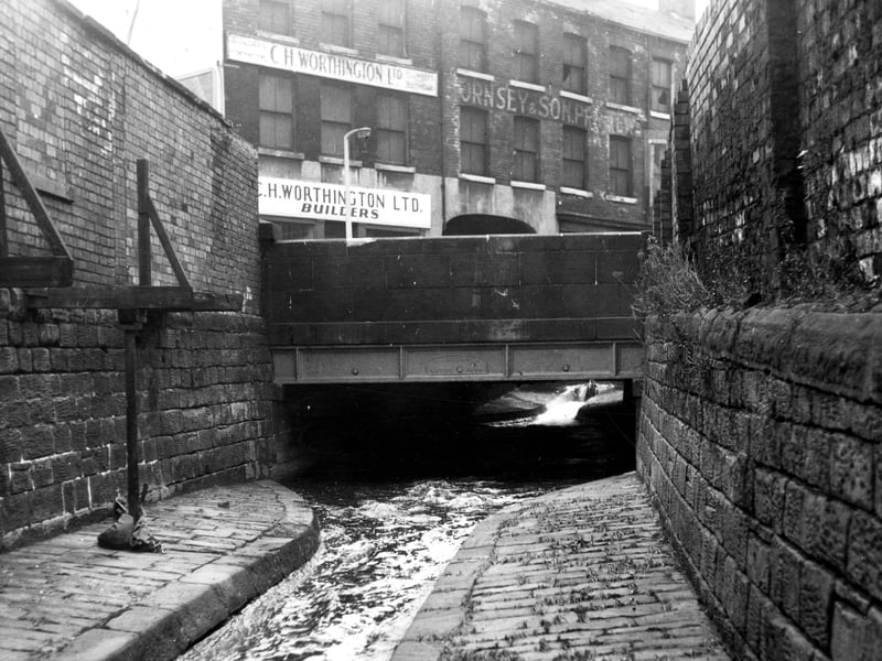 This view shows a footbridge over Low Beck by Holbeck Lane. Low Beck is a continuation of Hol Beck. In the background are the premises of C.H. Worthington Ltd., builders and contractors, and Hornsey & Son, printers, both of Holbeck Lane. Pictured in November 1965.