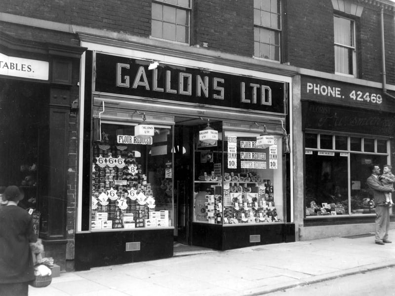 Gallons Ltd on Harrogate Road in September 1938. This is a grocers with a double fronted window showing wares. On the right is TW Smith Florist and a man holding a young child is stood on the pavement.