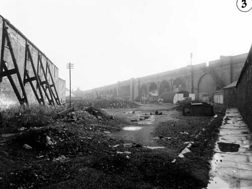 Wasteground in the Isle Lane area, showing the rail viaduct in the background. On the left a high brick wall is shored up with wooden struts. On the right a Nissen hut and a car can be seen. Pictured in November 1949.