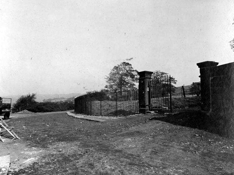 Fawcett Lane improvement pictured in September 1934. Stone gateposts with iron gates and railings, surrounding grassy area with trees. Waste ground in foreground. Distant view of houses and factory chimney.