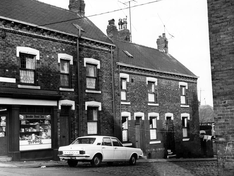 Early 1970s image shows on the left number 65 Elsworth Street, a corner shop grocery run by J. Hanley. Products advertised and viewed through the window include a variety of cigarettes, tobacco's and tea's. On the right are a row of blind back terraced houses with a shared outside toilet on the right. Clothes hang from lines stretched across the street while outside number 46 Winchester Street a Zephyr, reg: MNW 744F is parked.