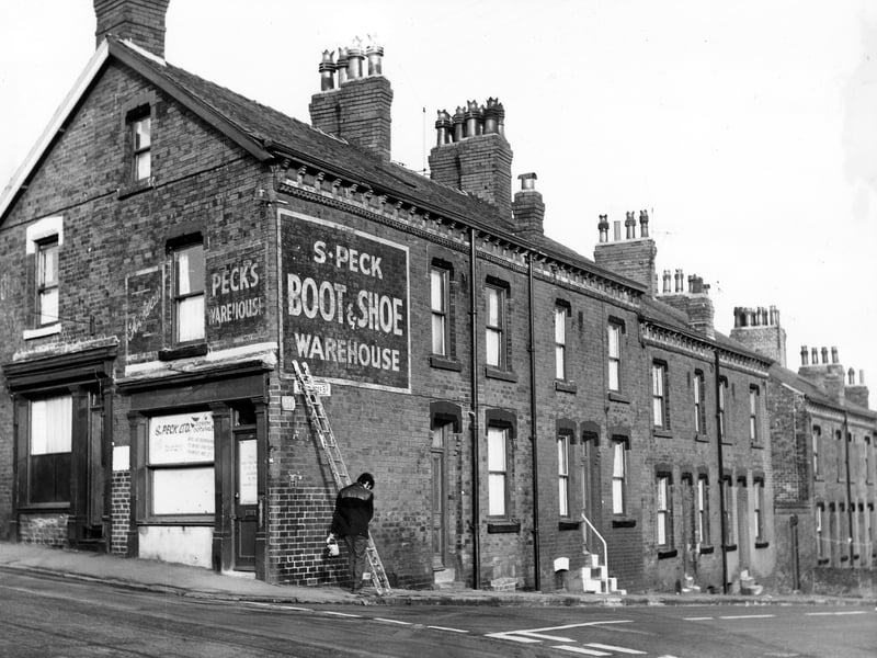 This early 1970s photo shows two shop fronts on the left numbers 26 and 28 Hall Lane. Number 26 on the right is advertised as the S. Peck Boot and Shoe Warehouse. A man wearing a donkey jacket holding a tin of paint and brush stands at the corner by a ladder. On the right are numbers 82 to 88 Winchester Road a row of back-to-back terraced houses. These houses are located opposite H.M. Prison Armley.