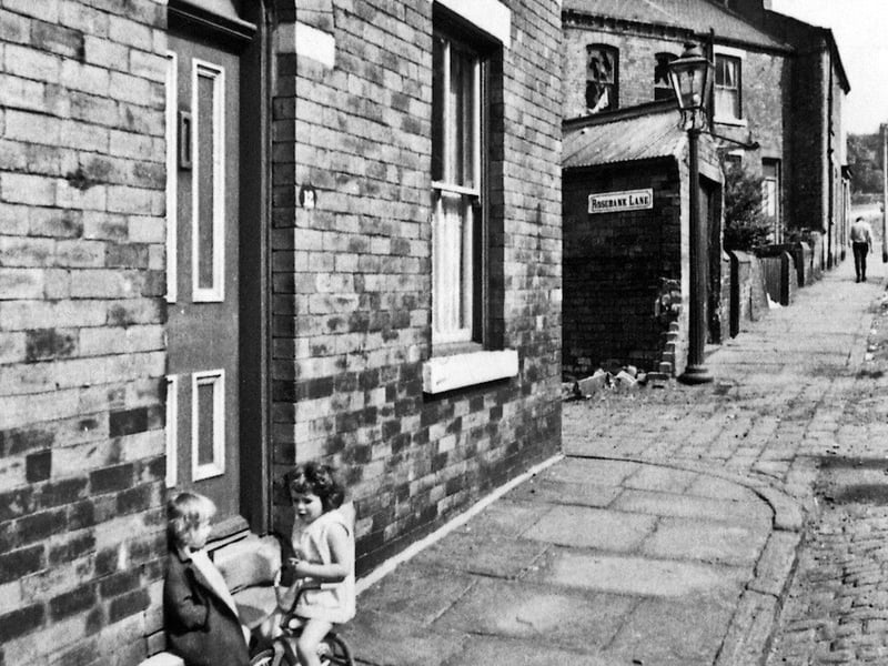 A small girl on a trike chats to another little girl seated on the doorstep of number 12 Westfield Crescent. The gas lamp stands at the narrow access to Rosebank Lane where the rear of the Sunday School and Congregational Church fronting Woodsley Road are located.