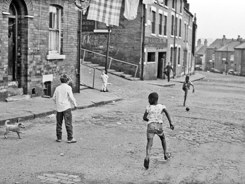A group of young boys play a game of football in Westfield Crescent while a toddler leaning against a gas street lamp looks on. Even the dog is joining in.