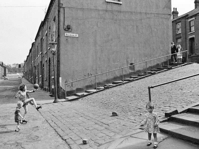 Children playing in Westfield Crescent, left, and Rillbank Street, right. A girl on roller skates practices her ball skills while a small boy watches. A little girl in the foreground looks directly at the photographer. Steps with handrails flank the cobbled Rillbank Street and a young man and a woman begin the descent from Back Rosebank Grove.
