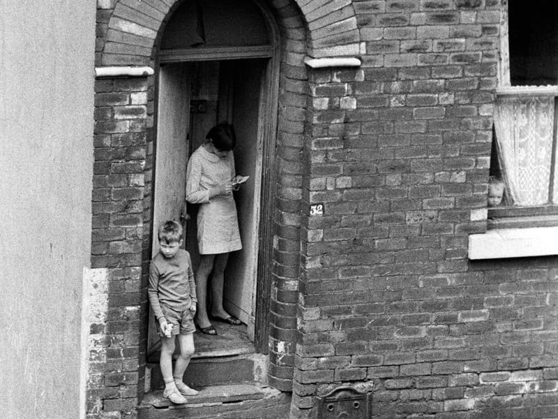 Rosebank Grove showing no. 32, situated at the end of a terrace, with the top of a flight of steps which lead down to Westfield Crescent just visible on the left. A young boy stands in the doorway, with a young woman just behind him reading a letter, while a smaller child can just be seen peeping out of the window and a dog is in the street outside the door.