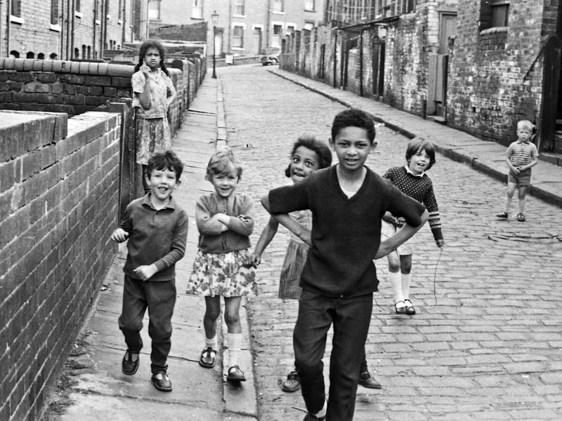 In this view of Back Rosebank Grove, a group of children playing out in the street pose smiling for the camera while a small boy and an older girl look on with uncertainty.
