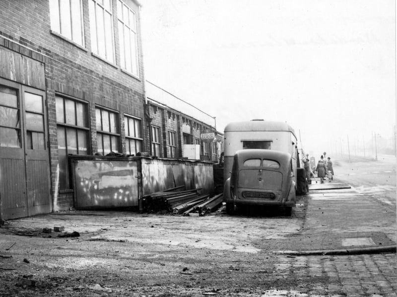 A car and a caravan outside Wandahome caravan sales at number 121 Stanningley Road. A policeman, two women and a child are by a belisha beacon. A pile of scrap metal and an advertisement for Redline petrol are visible. Another man appears to be cleaning the caravan. Pictured in November 1951.