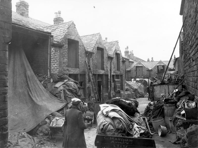 A yard off Winker Green. On the left and at the rear are small workshop/garage style buildings. The whole yard is filled with indusrial debris and scrap metal. In the foreground a man stands next to a overloaded wheel barrow with 'C. Hanshaw, Armley Leeds' printed on the side. Pictured in November 1957.