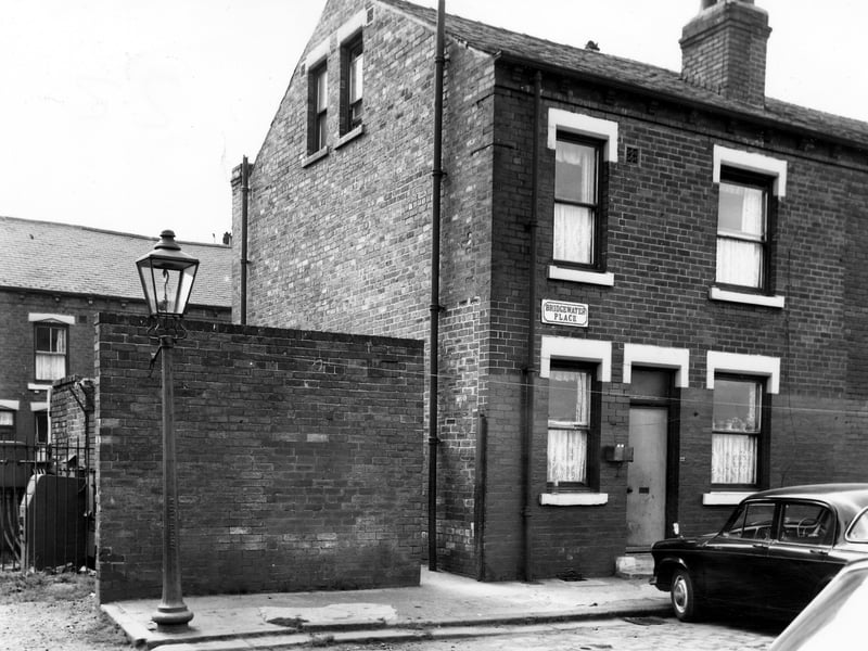 The start of a row of back-to-back terraced houses on Bridgewater Place pictured in July 1965. A box has been mounted on the left of number 22 for milk deliveries. On the left is a shared outside toilet block and ginnel leading through to Bridgewater Terrace. Visible on the far left is an electricity sub station surrounded by railings