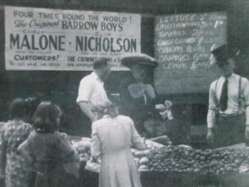 A fruit and vegetable stall at a garden party in the grounds of Mount St Mary's Church. The men behind the stall are Tommy Nicholson, Andrew Malone, and Tommy O'Hearne. The sign behind announces them as 'Curly' Malone and 'Slim' Nicholson.