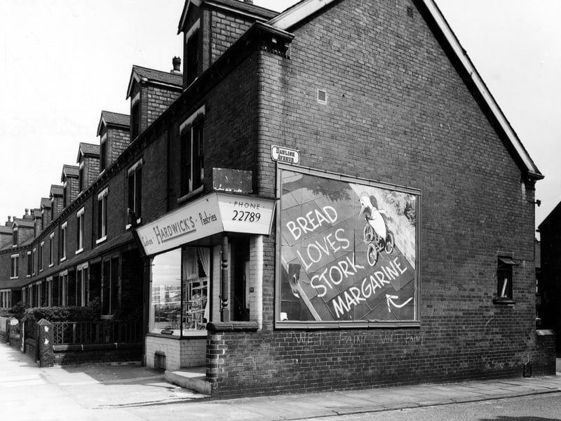 This view from May 1956 looks north at the junction with Dawlish Avenue. Hardwick's pastries and cakes is on end of row of three storey brick terraces with dormer windows. Large advertising sign for Stork margarine on gable end.