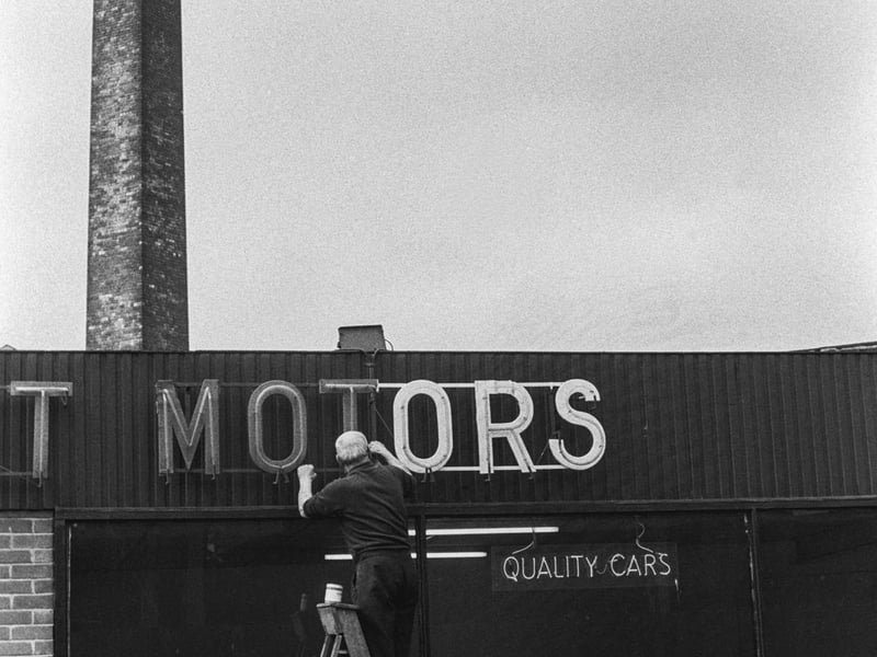 A man on step-ladders as he appears to attend to the sign above the windows of a garage. The garage is East Street Motors of number 28 East Street. A sign in the window advertises 'Quality Cars'. In the background, left, a large square chimney looms which is located in Waddington's Yard, between East Street and the River Aire.