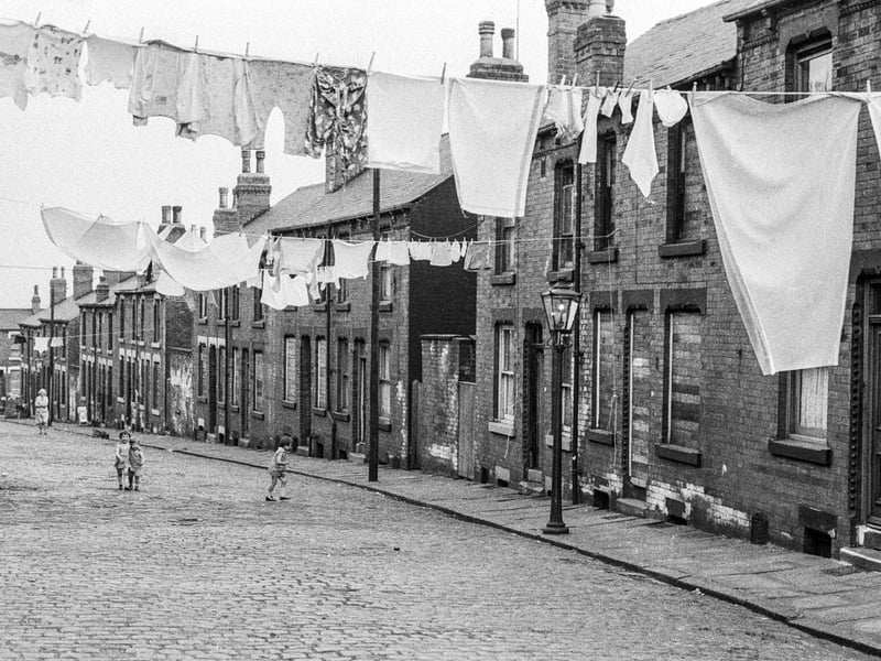 Terraced housing on Rosebank Grove, looking down towards Rosebank Mount on the far left. Children are playing on the road and lines of washing are hung across. No. 52 is on the right followed by no. 54 which has its door and windows bricked up. Yards containing shared outside toilets separate each block of four houses.
