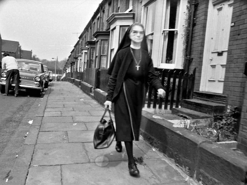 A nun glances towards the camera as she walks along Burley Lodge Road, past red brick terraced homes with bay windows and small walled gardens. On the other side, left, more terraced streets go off Burley Lodge Road to join Carberry Road, possibly Carberry Place and Back Carberry Place.