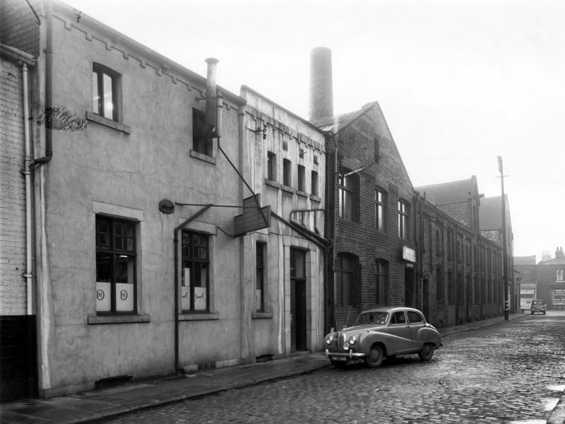 Admiral Street in December 1958. The white building on the left is number 8, the Davy Machine Co fish fryers engineers. In the window on the left has been etched the logo of an M inside a D while the windows on the right display the Triumph logo. A car, reg: MWY 666 is parked on the right is number 6 the Maple Clothing Co blouse factory, a sign hangs over the office entrance. In the distance can be seen Dewsbury Road.