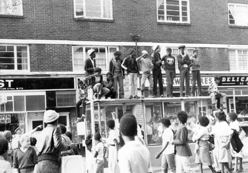 Crowds are gathered in Chapeltown Road on August Bank Holiday Monday in 1980 for the Leeds West Indian Carnival. A group of youngsters have claimed a higher vantage point on top of a bus shelter.