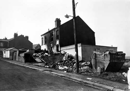 Sarah Street showing the rear of No.58, left and 60, right, Fall Lane. There are visible signs of a fire having occurred. Skips are adjacent to the premises for the collection of waste materials as the houses were about to be renovated. At the left edge is No.1 Sarah Street and a row of garages. The roof of St. Gabreil's Mission Church can be seen between the buildings.