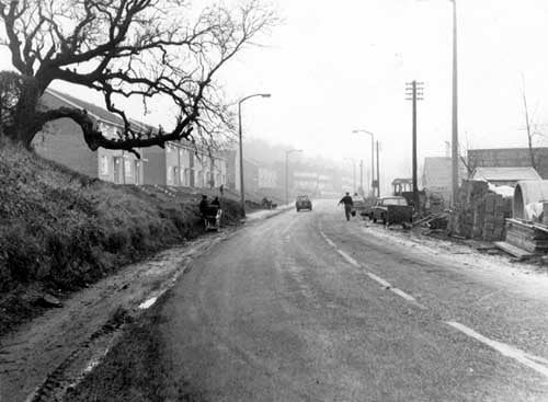 Pudsey Road, just east of the Ring Road. People, traffic and houses visible. Construction work to the right. Street lights and telephone poles visible. Pictured in December 1968.
