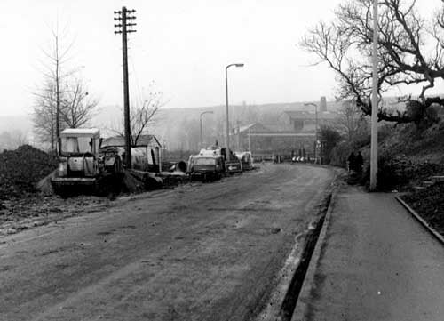 Looking west-north-west along Pudsey Road in December 1968. Roadworks and diversion signs visible, also parked cars and a bulldozer. Two women are walking along the street. A bus can be seen in the distance. Junction with Henconner Lane visible.