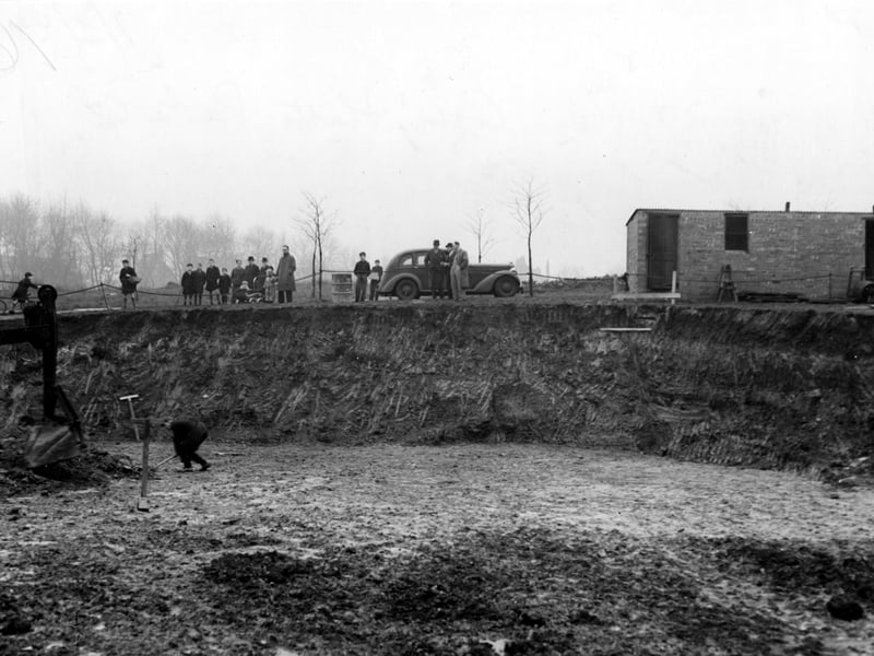 Showing excavations in Cross Flatts Park in November 1942. A workman stands in the large hole, where the front of a digger is visible. On the rim of the hole are members of the public and a parked car.