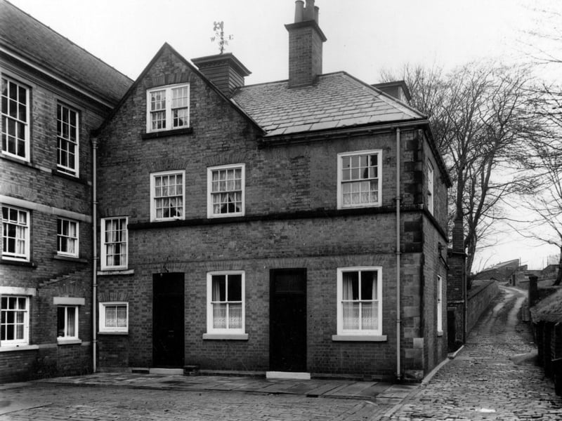 Photo of the rear of Allerton Hall with a stone set yard and path leading up hill. The brick annex is three storeys high with a gable and chimneys. There is a metal weather vane on top of roof. Pictured in November 1950.