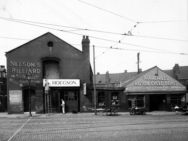 The west side of Harehills Road showing Nelson's Billiard Hall, Hodgson's and Harehills Motorcycle Depot with a young boy looking in the window and a motorcycle outside. Man on ladder painting sign. Pictured in August 1947.