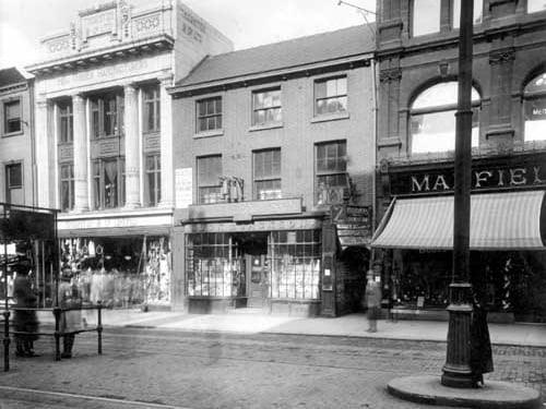 Briggate with Wm Green and Son, grocers to centre of the photo from June 1922. Turks yard is to right with signs for Whitelocks luncheon bar clearly visible. Also showing is Manfield and Sons Boot dealers. To left of Greens is Thorton and Co. Ltd. India Rubber manufacturers. Two children wait in the tram stop.