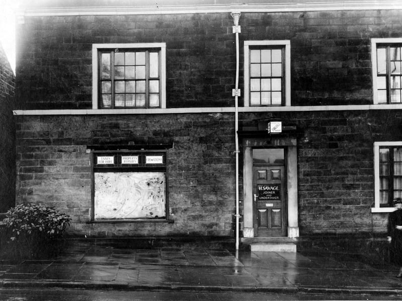 Detached stone property on Harrogate Road, the business of Hubert Savage. Lamp in doorway and sign on the door inform 'Joiner and Undertaker'. The window on the left has been white washed, panels to the top have advertisments 'Taxis for hire, Property repairs, Cremations' Pictured in  December 1936.